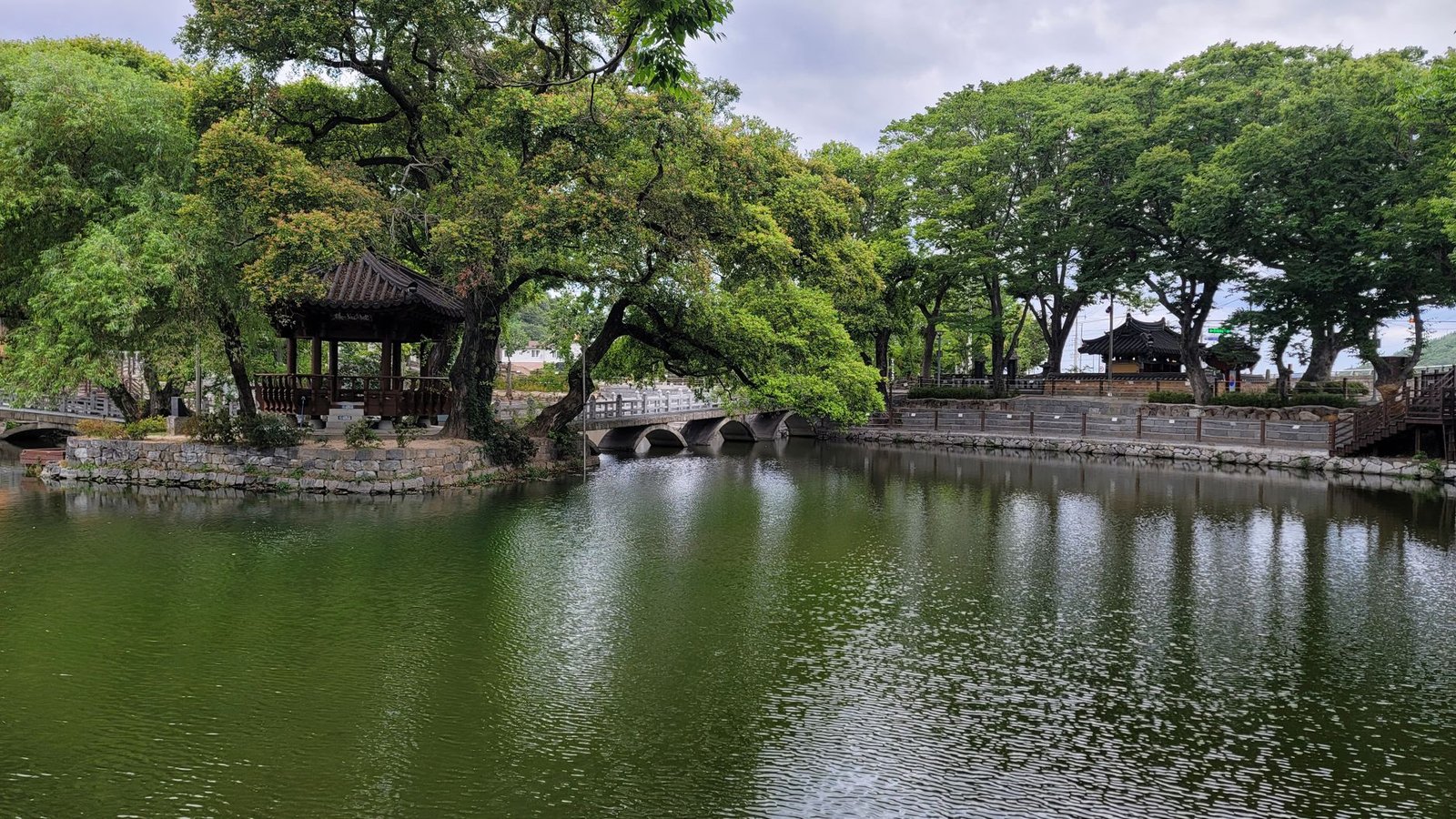 A tranquil park scene featuring a traditional pavilion, bridge, and lush greenery reflected on a calm pond