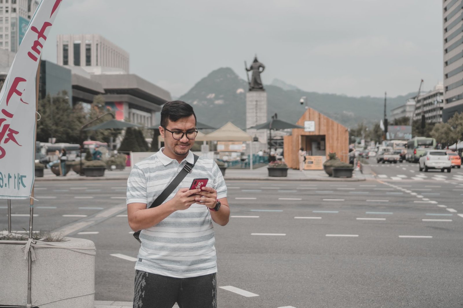 A man in a striped shirt standing on a street using a smartphone with a city skyline in the background